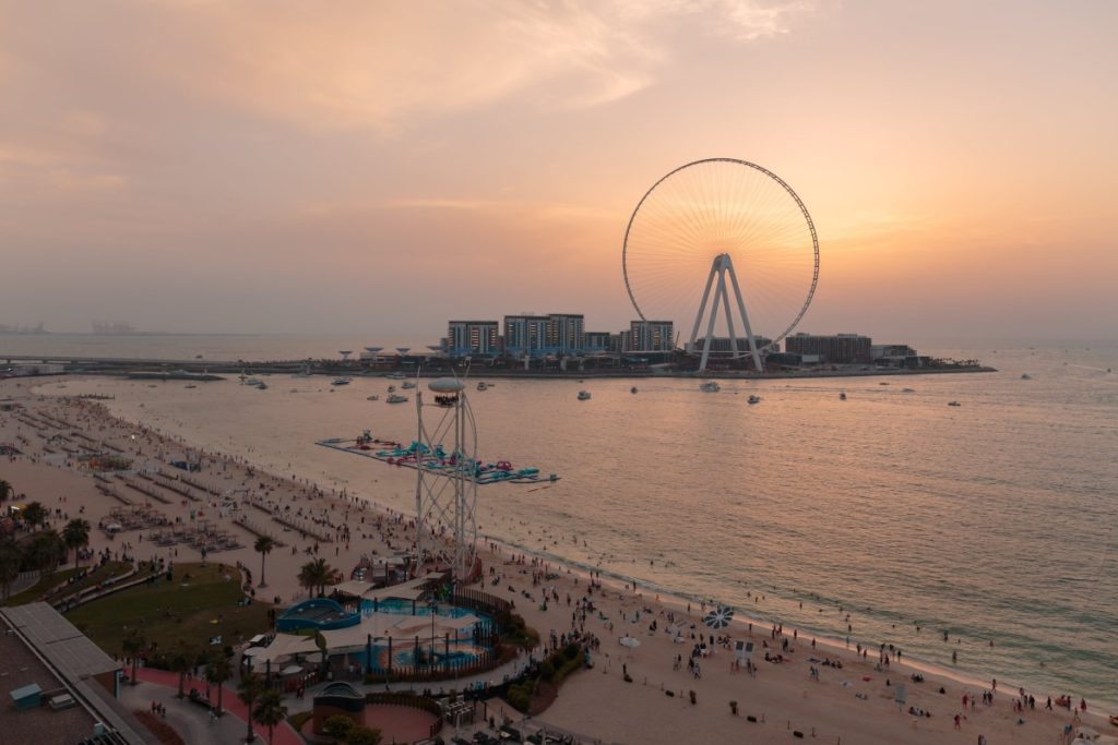 A group of people seated in circular booths attached to a rotating platform high in the sky, overlooking the cityscape of Dubai. Each booth is equipped with safety harnesses, providing a thrilling dining experience as guests enjoy food and beverages while taking in panoramic views from above.