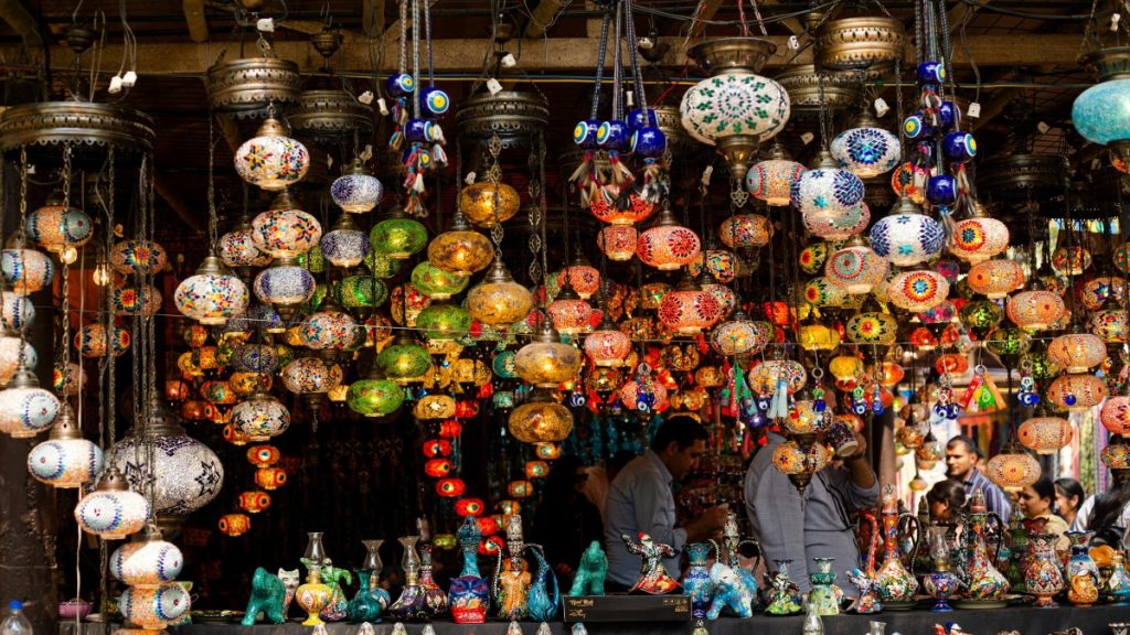 A bustling scene within the Istanbul Grand Bazaar, showcasing colorful arrays of textiles, vibrant spices, and gleaming lanterns, as shoppers and vendors navigate through the maze of narrow alleys, immersing in the vibrant atmosphere of this historic marketplace.